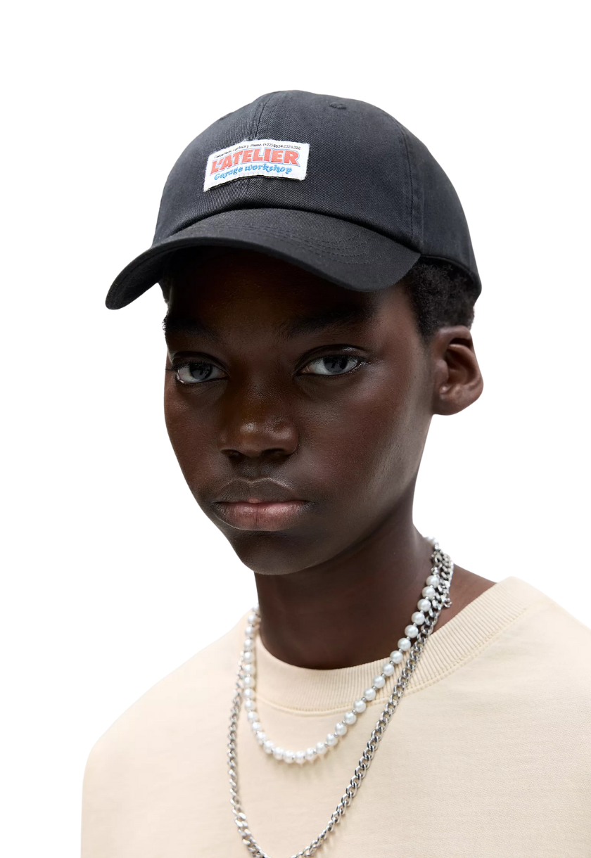 Gorra negra estilo mecánico para hombre con la palabra “L’atelier” bordada en negro y ubicada encima de un cuadrado blanco. Incluye el modelo